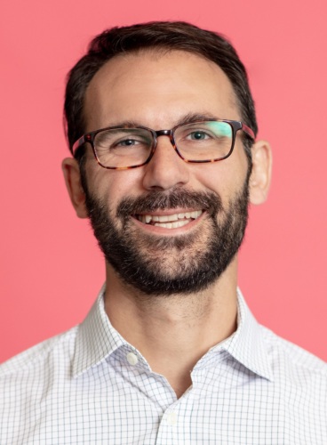 Jake Edwards, a man with short dark hair, glasses, and a beard, is smiling against a pink background. He is wearing a white shirt with a small checked pattern.